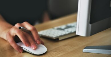 A hand using a wireless mouse at a modern desk setup with a computer and keyboard.